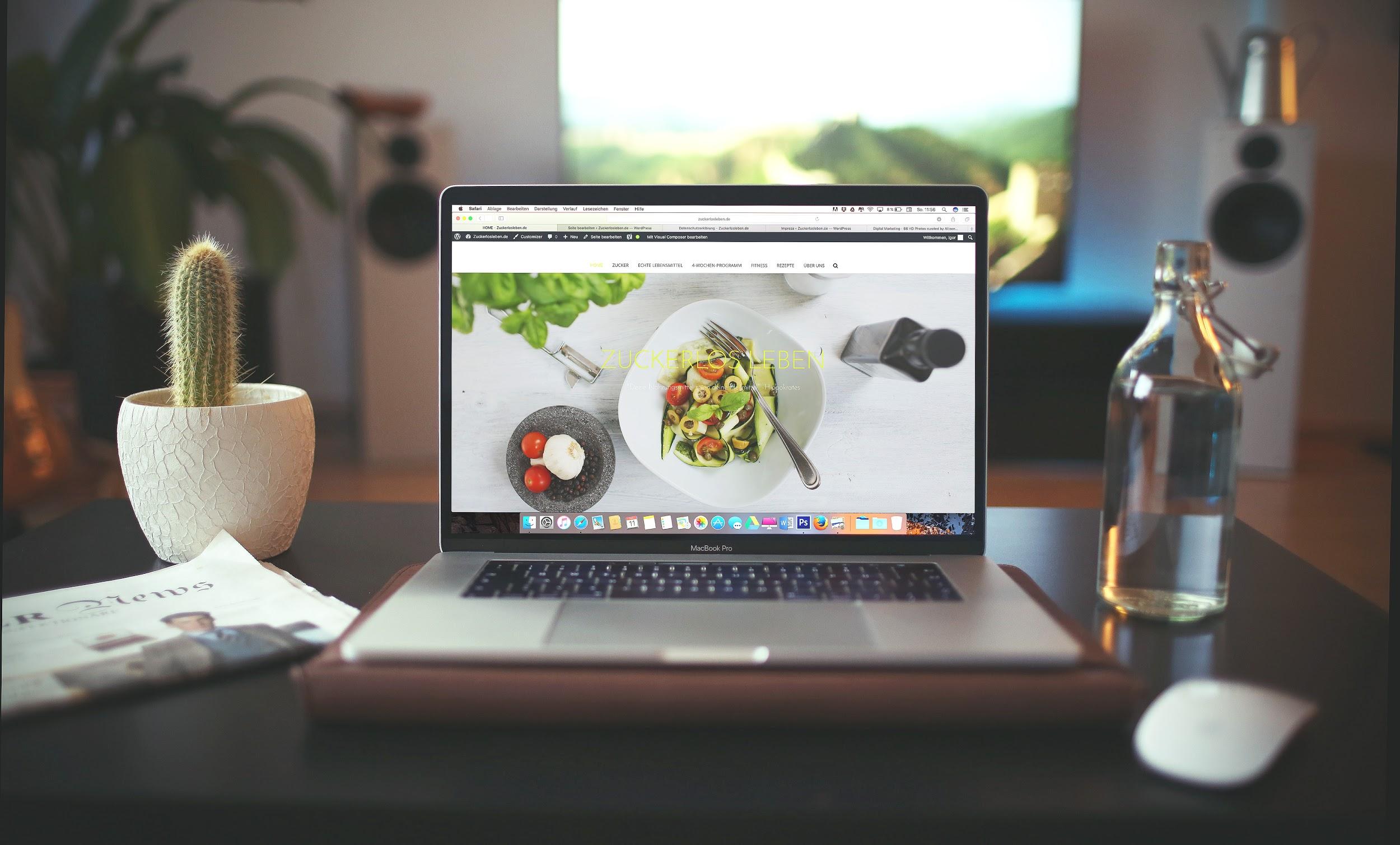 A laptop sits open on a desk showing a website with a bowl of vegetables. The desk also contains a plant, a newspaper, and a glass of water.