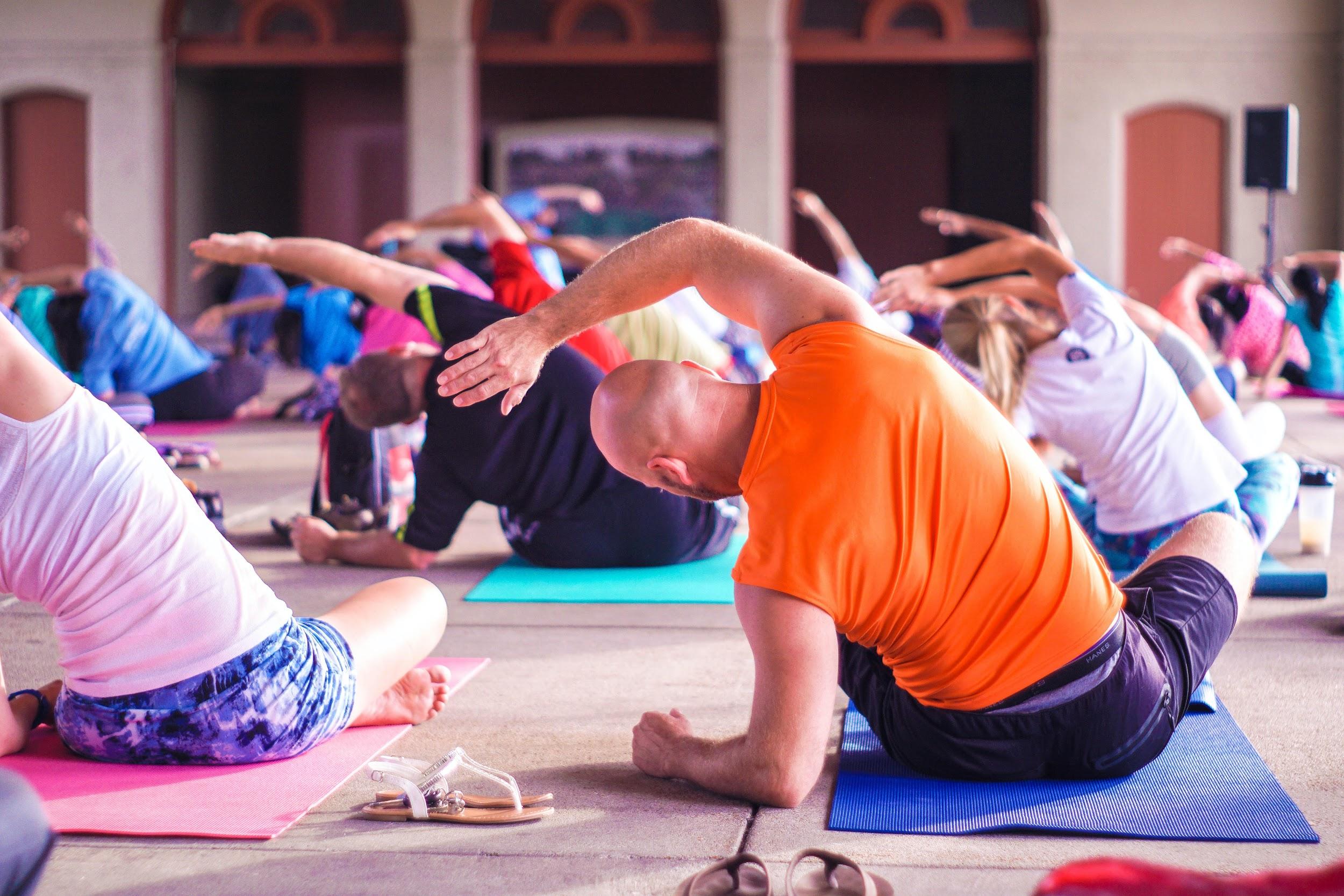 A group of people are shown sitting on yoga mats and participating in a stretching exercise.