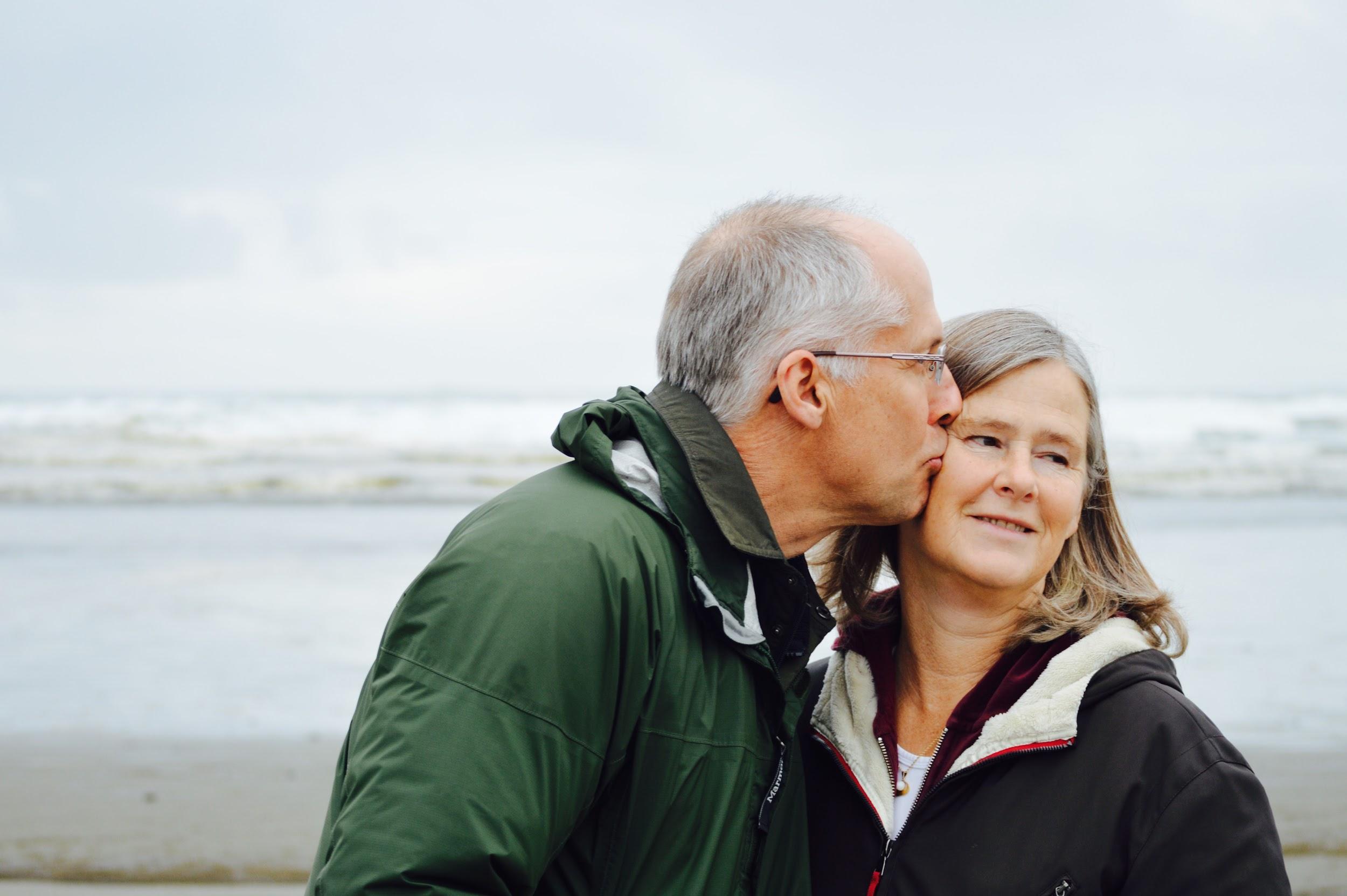An older male and older female stand on the beach with the ocean behind them. The man is kissing the woman on the cheek.