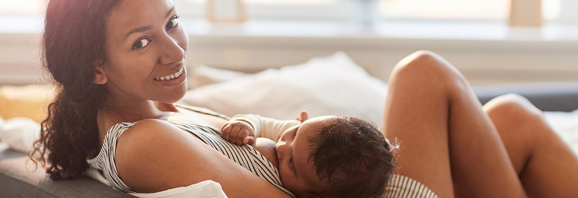 A woman with darker skin, and brown curly hair sitting on the couch smiling and breastfeeding her baby.