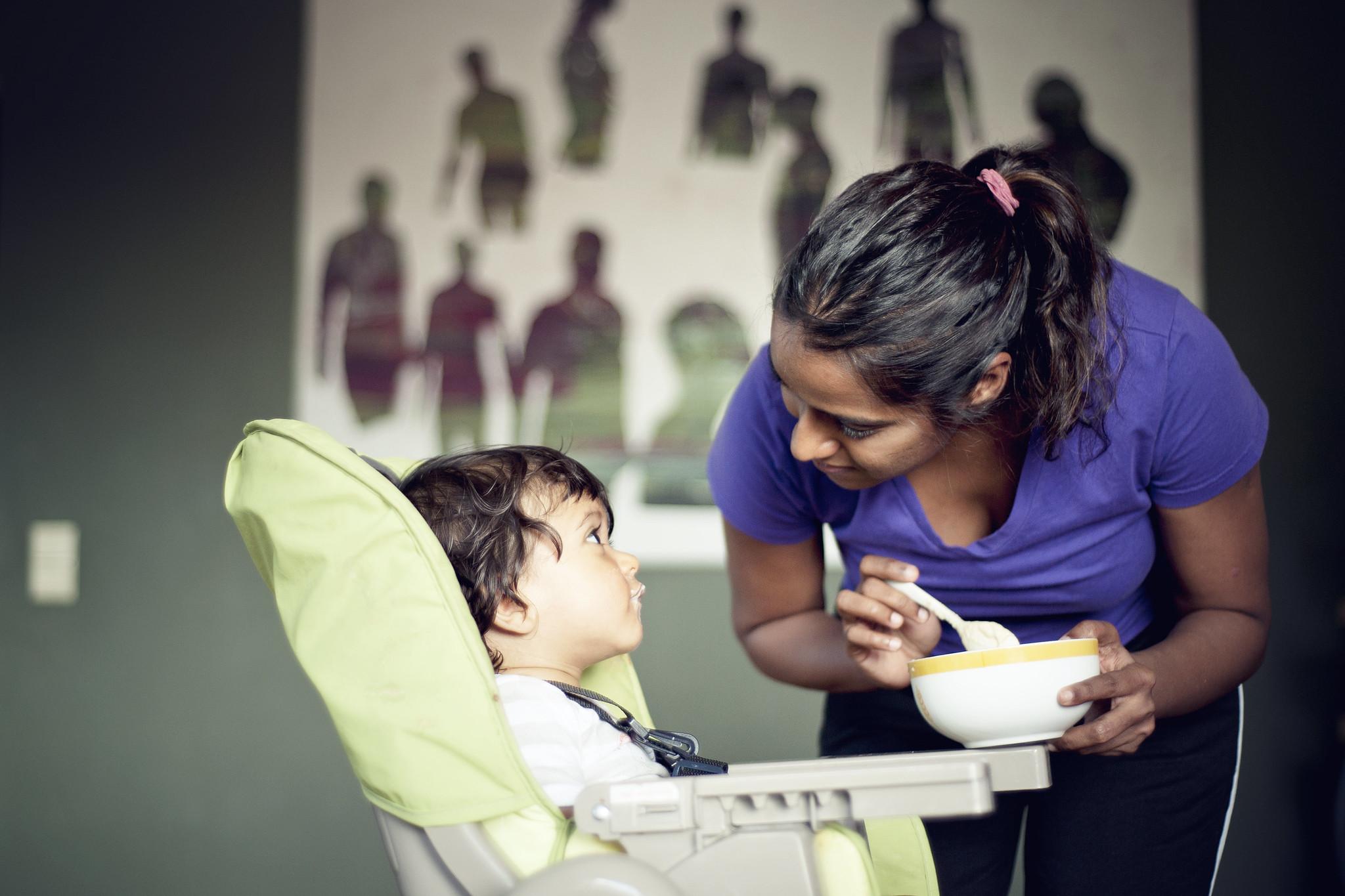 A mother with brown skin tone makes eye contact with a baby in a high chair. The mother is holding a bowl of baby food and a spoon and looks like she's pausing before offering another bite of food to the baby.