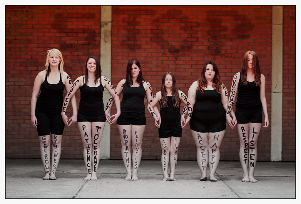 Six young women stand in a row wearing black leotards. The women are of varying body shape and size. They have positive phrases written in large letters on their arms and legs, such as patience, tolerance, be supportive, and be positive.