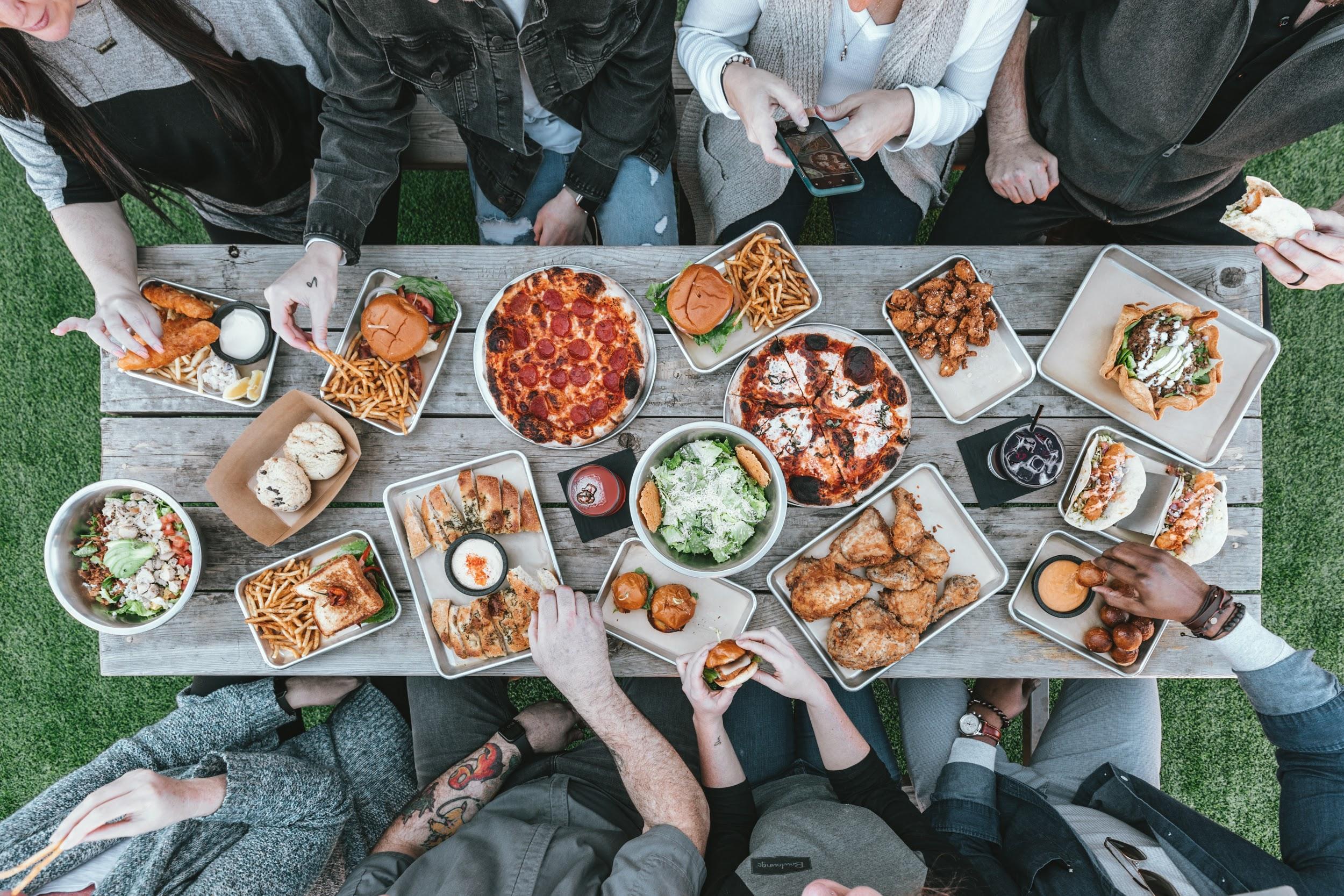 The photo shows a wooden picnic table surrounded by people (just arms and legs showing). On the table are trays holding a variety of foods, including pizza, salads, sandwiches, tacos, and chicken.