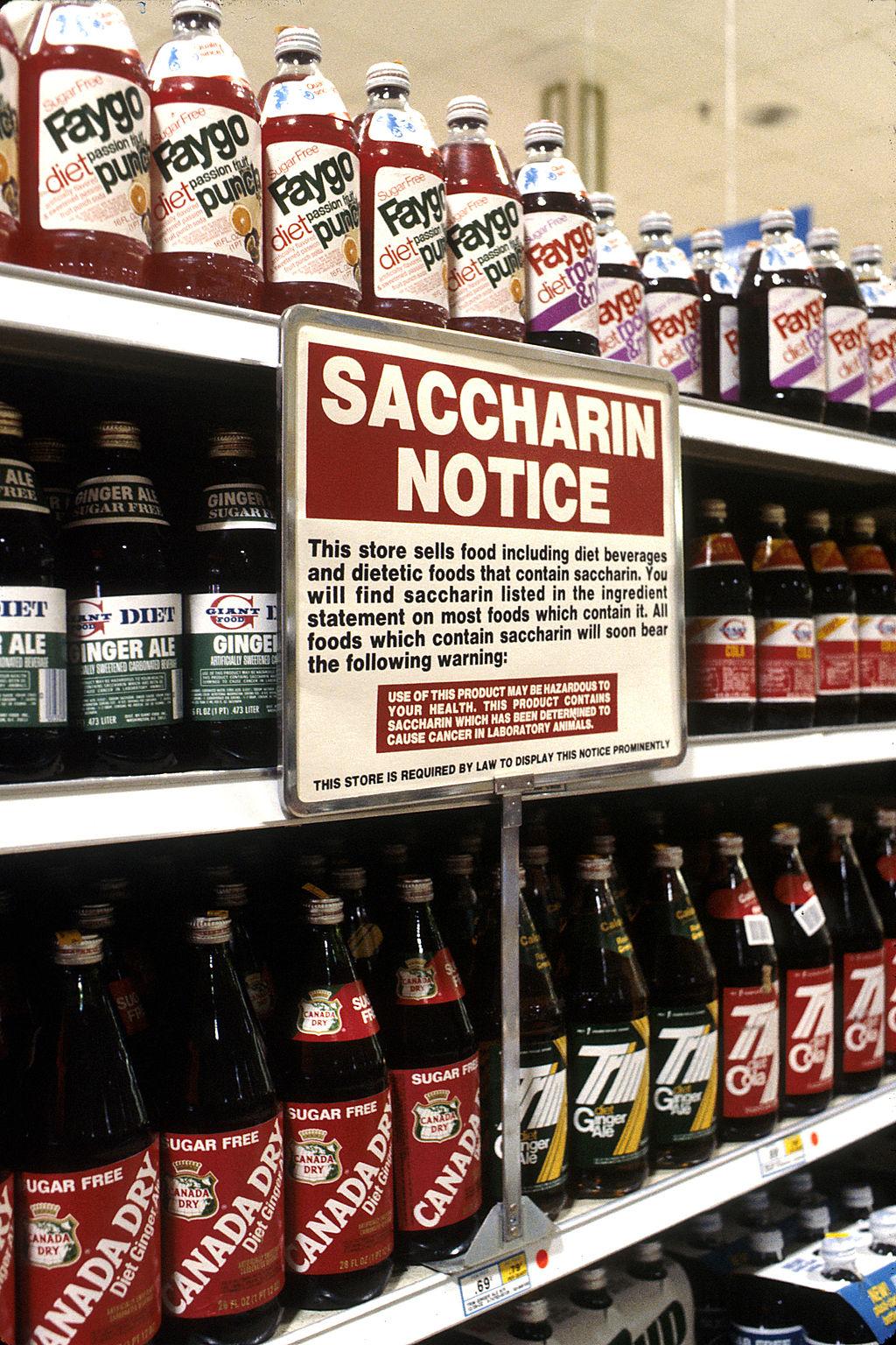 A photo shows a large sign titled "Saccharin Notice" posted in front of a shelf full of soda. Photo appears to be from the 1980's. The sign reads: "This store sells food including diet beverages and dietetic foods that contain saccharin. You will find saccharin listed in the ingredient statement on most food which contain it. All foods which contain saccharin will soon bear the following warning: Use the this product may be hazardous to your health. This product contains saccharin which has been determined to cause cancer in lab animals."
