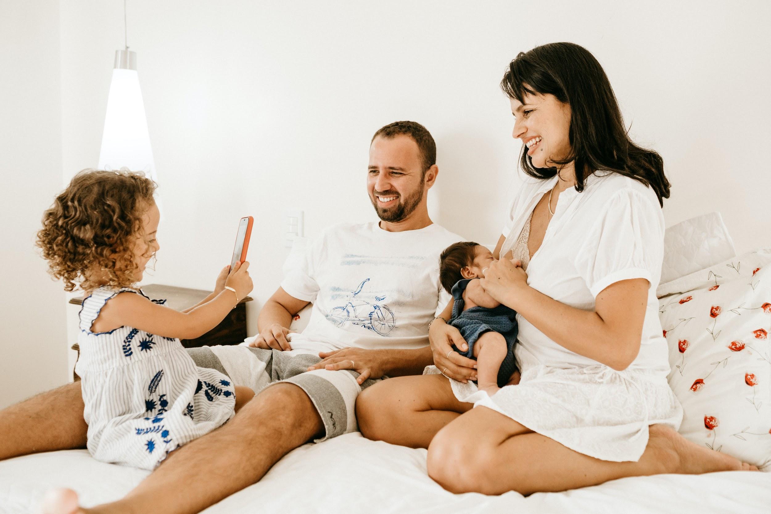 A family of 4 sits together on a bed. The mother is breastfeeding a young infant, and a young child with curly hair appears to be taking a picture of them with a phone. The father smiles and looks on.