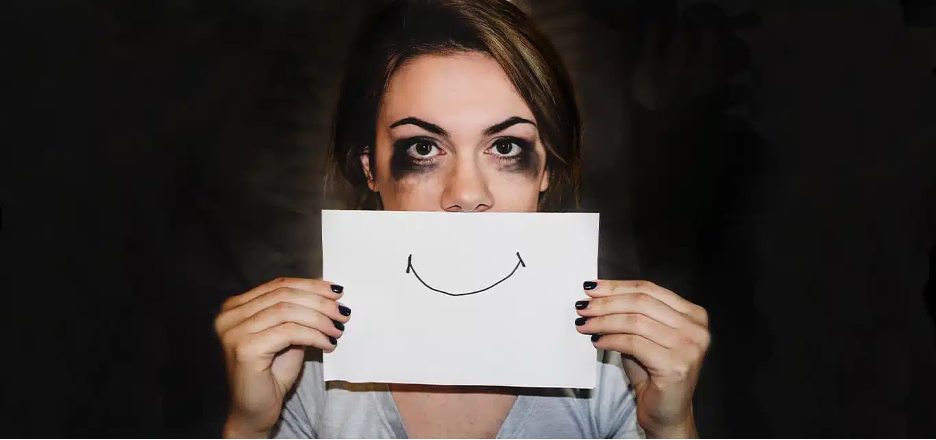 A distressed woman with smudged mascara holds up a paper with a drawn smile, hiding her true emotions.